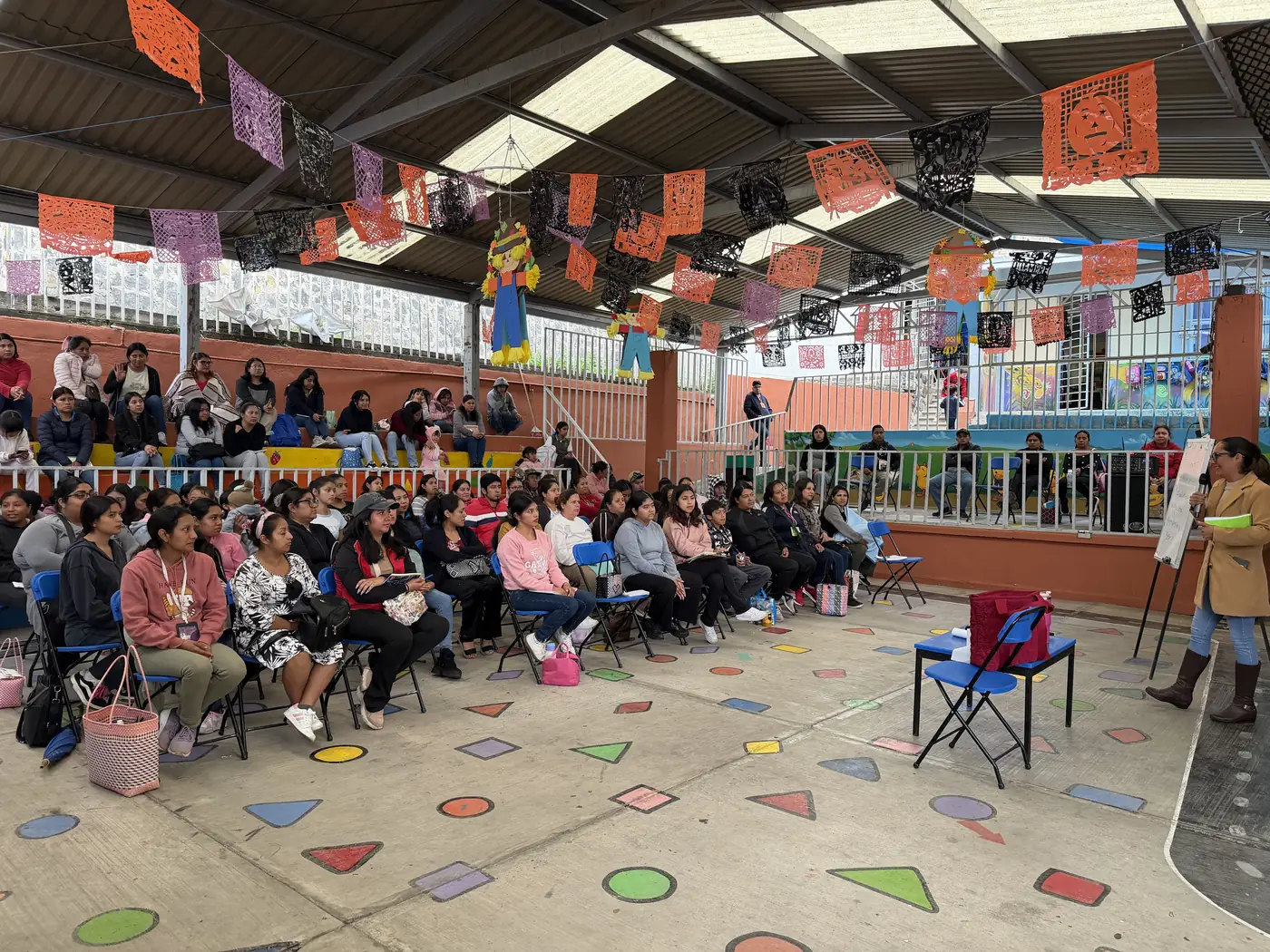 Taller comunitario con madres, padres y familias escuchando a una facilitadora en un patio escolar.